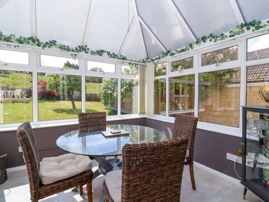 A conservatory with a round glass table and wicker chairs overlooking a garden at Hillside in Bishops Cleeve
