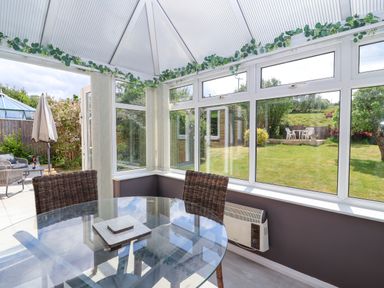 A conservatory with a glass dining table wicker chairs and a view of a garden at Hillside in Bishops Cleeve