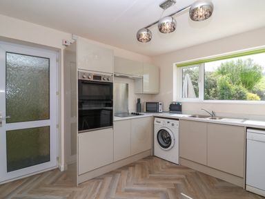 A kitchen with built-in oven, washing machine, microwave, sink, and cabinets with a window showing greenery at Hillside in Bishops Cleeve