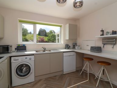 A kitchen with a washing machine, dishwasher, sink, microwave, toaster, kettle, counter stools, and a window with a view of trees and grass at Hillside in Bishops Cleeve