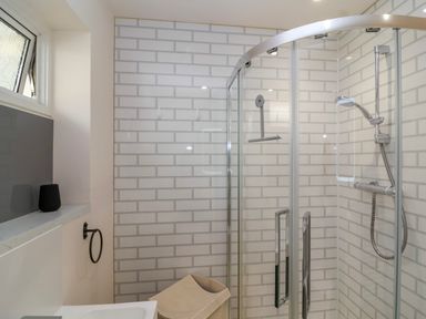 A bathroom with a glass shower enclosure white brick wall tiles a window and a laundry basket at Hillside in Bishops Cleeve