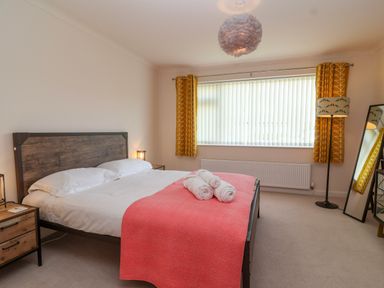 A bedroom with a double bed with white linens and a coral blanket surrounded by bedside tables and lamps with yellow curtains on the window at Hillside in Bishops Cleeve
