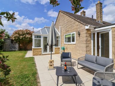 A patio with outdoor furniture and an umbrella next to a stone house with a conservatory and garden at Hillside in Bishops Cleeve