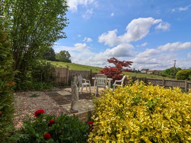A garden patio area with white plastic chairs and table a stone statue and yellow and red plants at Hillside in Bishops Cleeve