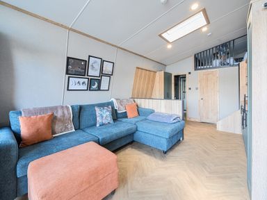 A living room with a blue sectional sofa orange ottoman wooden floor and framed pictures on the wall at Starlight Skylight - Billing Aquadrome in Billing Aquadrome Holiday Park