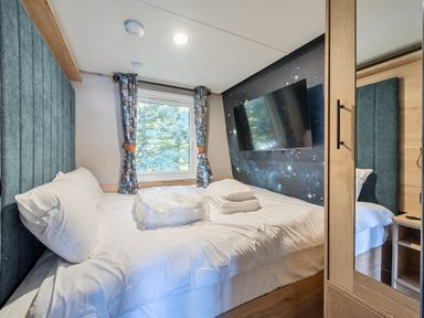 A bedroom with a bed, pillows, folded towels, a window with curtains, a wall-mounted TV, and a wardrobe with a mirror at Starlight Skylight - Billing Aquadrome Holiday Park