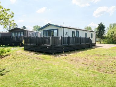 Two single story holiday homes with dark railings on raised wooden decks surrounded by grass and trees at Aquadrome Abode - Billing Aquadrome in Billing Aquadrome Holiday Park