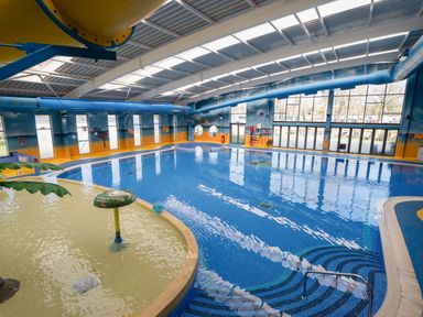 An indoor swimming pool with blue water a shallow play area with water features and large windows at Lakeside Lullaby - Billing Aquadrome in Billing Aquadrome Holiday Park