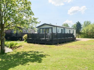 Two mobile homes with black wooden decks and railings on a grassy area with trees at Lakeside Lullaby Billing Aquadrome