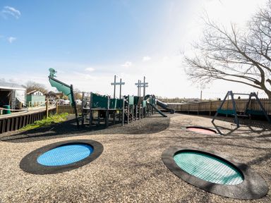An outdoor playground with a ship-shaped climbing structure slides swings and three in-ground trampolines at Moonlit Skylight - Billing Aquadrome in Billing Aquadrome Holiday Park