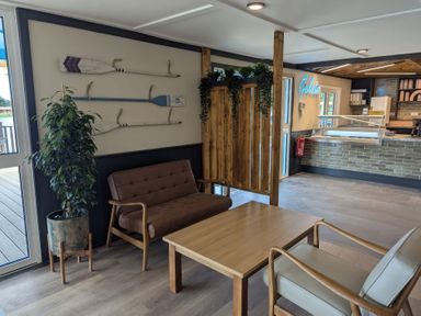 A seating area with a brown sofa, a wooden table, a beige armchair, a potted plant, wall-mounted oars, and a gelato counter at Moonlit Skylight - Billing Aquadrome in Billing Aquadrome Holiday Park