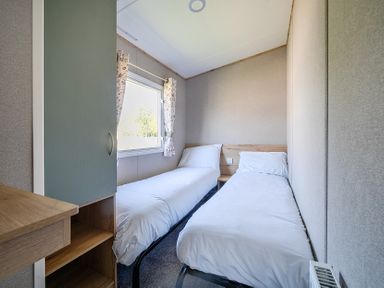 A small bedroom with two single beds a window with curtains a wardrobe and a radiator at Willowbrook Hideaway - Billing Aquadrome Holiday Park