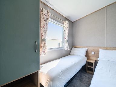 A bedroom with two single beds a small wooden nightstand and a window with patterned curtains at Willowbrook Hideaway - Billing Aquadrome Holiday Park