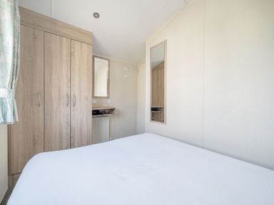A bedroom with a bed a wooden wardrobe two mirrors a small shelf and a window with patterned curtains at Golden Dune - Hayling Island in Hayling Island Holiday Park