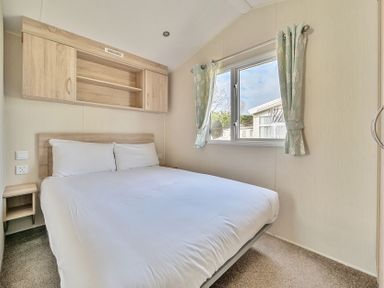 A bedroom with a bed a window with curtains and overhead cabinets at Golden Dune - Hayling Island in Hayling Island Holiday Park