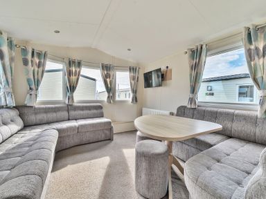 A sitting area with gray sofas a wooden table a small gray stool and windows with curtains at Golden Dune - Hayling Island in Hayling Island Holiday Park