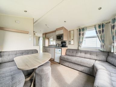 An open living area with a wooden dining table surrounded by grey upholstered seating a kitchen with cabinets a fridge microwave and appliances and windows with curtained at The Lookout - Hayling Island in Hayling Island Holiday Park