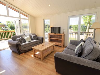 A living room with two gray sofas a wooden coffee table a wooden TV stand and large windows at Kingfisher Lodge in Gargrave