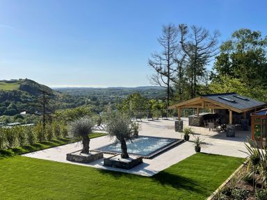 An outdoor area with a pool two trees on stone bases a covered seating area and green lawn at Forest View in Pontyclun
