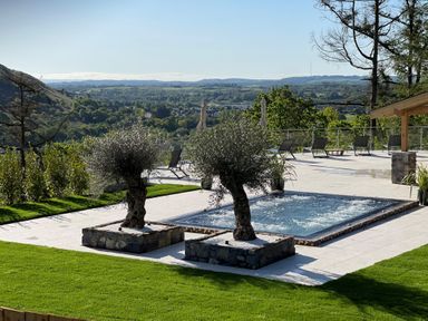 An outdoor area with two trees in stone planters next to a rectangular hot tub and several lounge chairs with umbrellas overlooking a valley at Forest View in Pontyclun