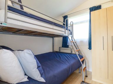 A bedroom with a metal bunk bed with blue bedding and a wooden wardrobe near a window with blue curtains at 101 Atlantic Bays Holiday Park in Padstow