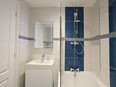 A bathroom with a white sink, a mirror with lights, a bathtub, and a shower with blue and white tiles at 10 Dartmouth House in Dartmouth