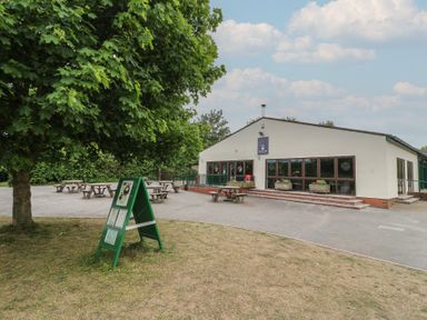 An outdoor cafe area with picnic tables and a green signboard near a building with large windows at Sycamore Lodge in Rosliston