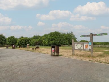 An outdoor park area with a paved path wooden trash bins information boards and directional signs with trees in the background at Sycamore Lodge in Rosliston