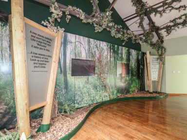An indoor room with wooden floor a forest mural on the wall large wooden signs with text and decorative branches hanging from the ceiling at Sycamore Lodge in Rosliston