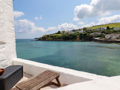 A balcony with a wooden table and storage box overlooking a coastal view with houses on a hill at Rock Cottage Portmellon near Mevagissey