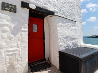 A red door set in a white stone wall next to a black outdoor storage box by the sea at Rock Cottage Portmellon near Mevagissey