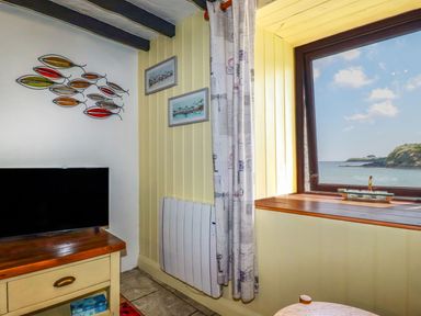 A corner of a room with a television on a wooden table metal fish wall art two framed pictures a window overlooking the sea and a curtain at Rock Cottage Portmellon near Mevagissey