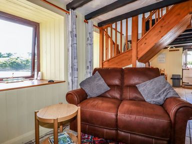 A sitting area with a brown leather sofa and two cushions next to a small wooden table and a window at Rock Cottage in Portmellon near Mevagissey