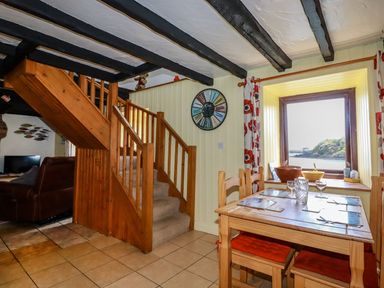 A dining area with a wooden table and chairs next to a window showing a coastal view and a wooden staircase in a living space at Rock Cottage in Portmellon near Mevagissey