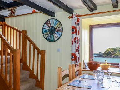 A dining area with a wooden table and chairs next to a window with a sea view and a staircase with wooden railing at Rock Cottage in Portmellon near Mevagissey