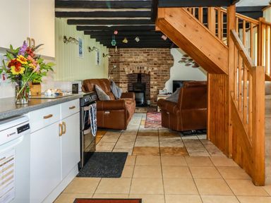 A kitchen with white cabinets and a countertop next to a living room with brown leather sofas and a brick fireplace at Rock Cottage Portmellon near Mevagissey