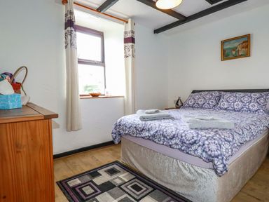 A bedroom with a double bed with patterned bedding a wooden dresser a window with curtains and a small rug on the floor at Rock Cottage Portmellon near Mevagissey