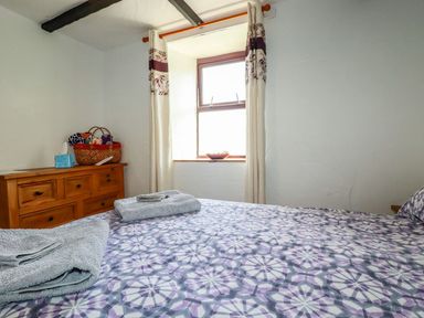 A bedroom with a patterned bedspread two folded towels on the bed a wooden chest of drawers with a basket on top and a window with curtains at Rock Cottage Portmellon near Mevagissey