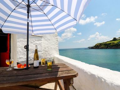 A wooden picnic table with drinks and fruit under a striped umbrella on a balcony overlooking the sea at Rock Cottage in Portmellon near Mevagissey