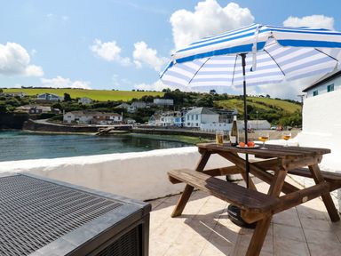 A wooden picnic table with drinks fruit and a bottle under a striped umbrella on a tiled patio overlooking water and houses at Rock Cottage in Portmellon near Mevagissey