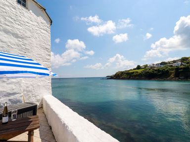 A seaside view from a balcony with a wooden table a striped blue and white umbrella and a white wall at Rock Cottage in Portmellon near Mevagissey