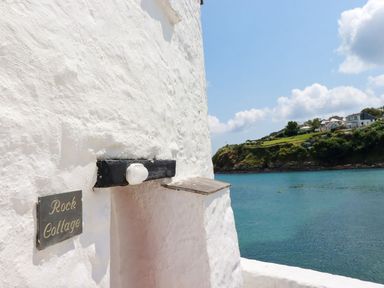 The side of a white building with a sign reading Rock Cottage overlooking a body of water with a grassy hillside and houses in the distance at Rock Cottage in Portmellon near Mevagissey