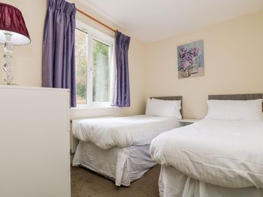 A bedroom with two single beds beside a window with purple curtains and a lamp on a white dresser at Valley Lodge 31 in St Ann's Chapel Cornwall