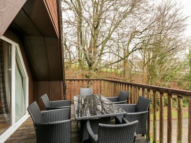 An outdoor wooden balcony with a black woven dining table and six matching chairs surrounded by leafless trees at Valley Lodge 31 St Ann's Chapel Cornwall