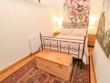 A bedroom with a metal bed frame a wooden chest at the foot of the bed two bedside tables with lamps and floral paintings on the walls at Shaftoe Mews in Haydon Bridge
