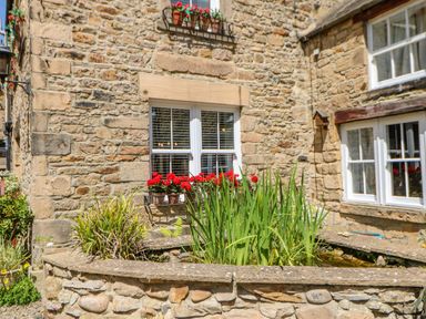 A stone wall with two white windows and flower pots with red flowers outside at Shaftoe Mews in Haydon Bridge