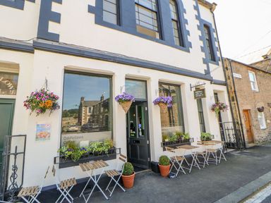 A cafe exterior with tables chairs and hanging flower baskets at Oddfellows in Shaftoe Mews Haydon Bridge