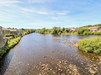 A river with stone houses and greenery on both banks at Shaftoe Mews in Haydon Bridge