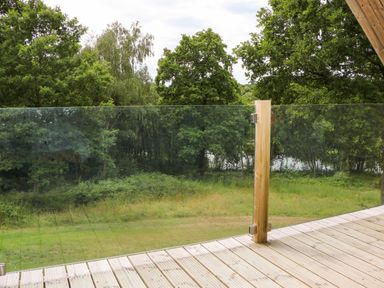 A wooden deck with a glass railing overlooking a grassy area and trees at Lakeside Lodge near Burwash