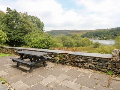 A picnic table and benches with a stone wall by a lake at Ty Newydd Llanrhychwyn near Llanrwst